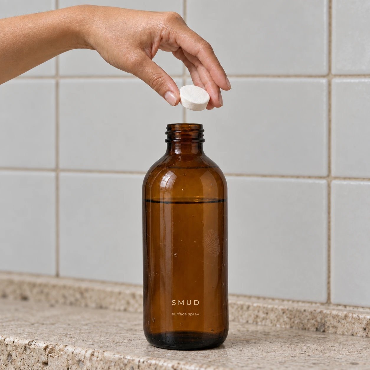 A hand dropping a refill tablet into a tall amber SMUD surface spray bottle filled with water